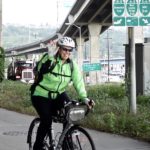 photo of woman riding bike on trail approaching Spokane Street Bridge. Semi truck and high bridge in background.