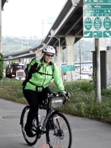 photo of woman riding bike on trail approaching Spokane Street Bridge. Semi truck and high bridge in background.