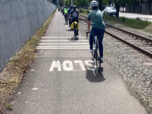 Group of bike riders on paved trail by railroad tracks and street.